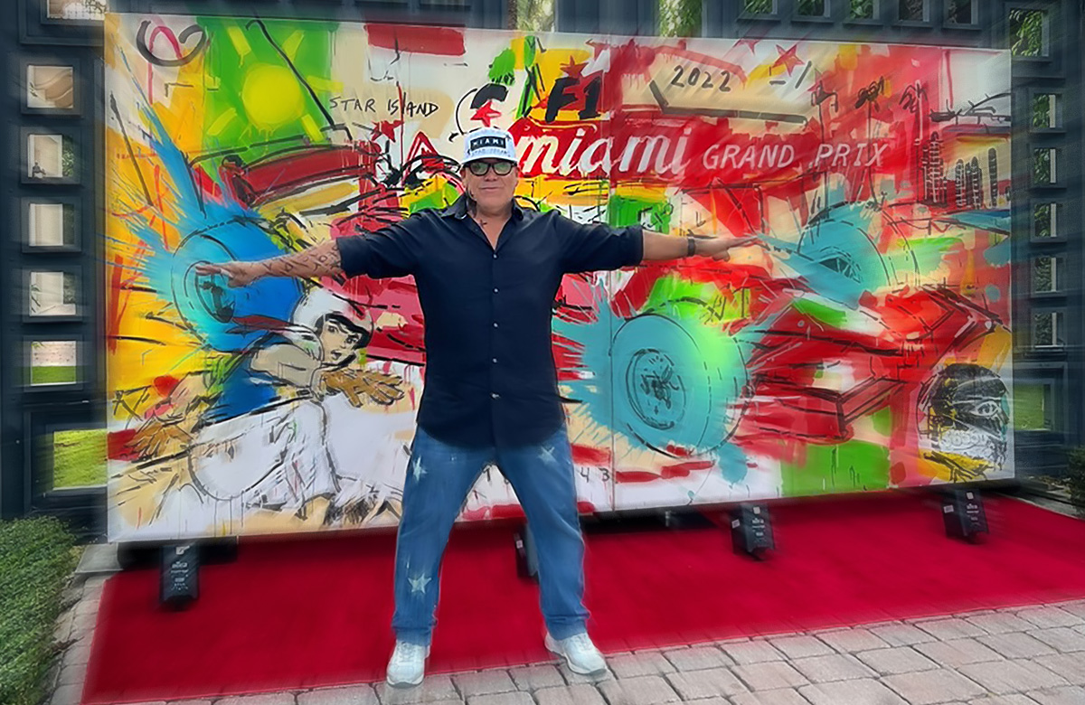 painter leonardo hidalgo in a dark blue shirt, blue jeans, white sport shoes and white baseball cap, standing with arms wide open in front of a big paint about racing cars